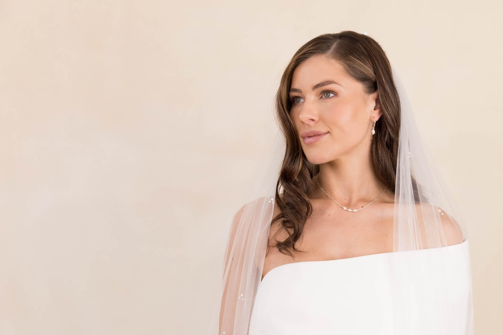 Woman wearing a white wedding dress with a bridal veil against a beige background.